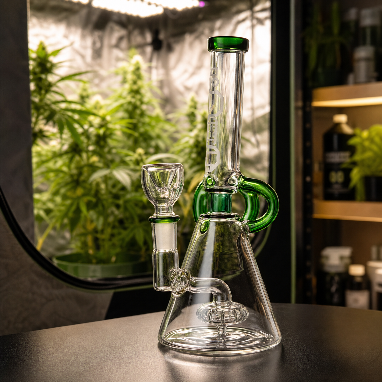 Bong de vidrio transparente con detalles verdes, base cónica y percolador visible, sobre una mesa frente a un cultivo interior.