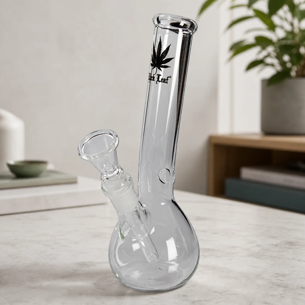 Bong transparente con base redondeada, tubo largo con una hoja negra y cazoleta lateral inclinada, sobre una mesa clara.