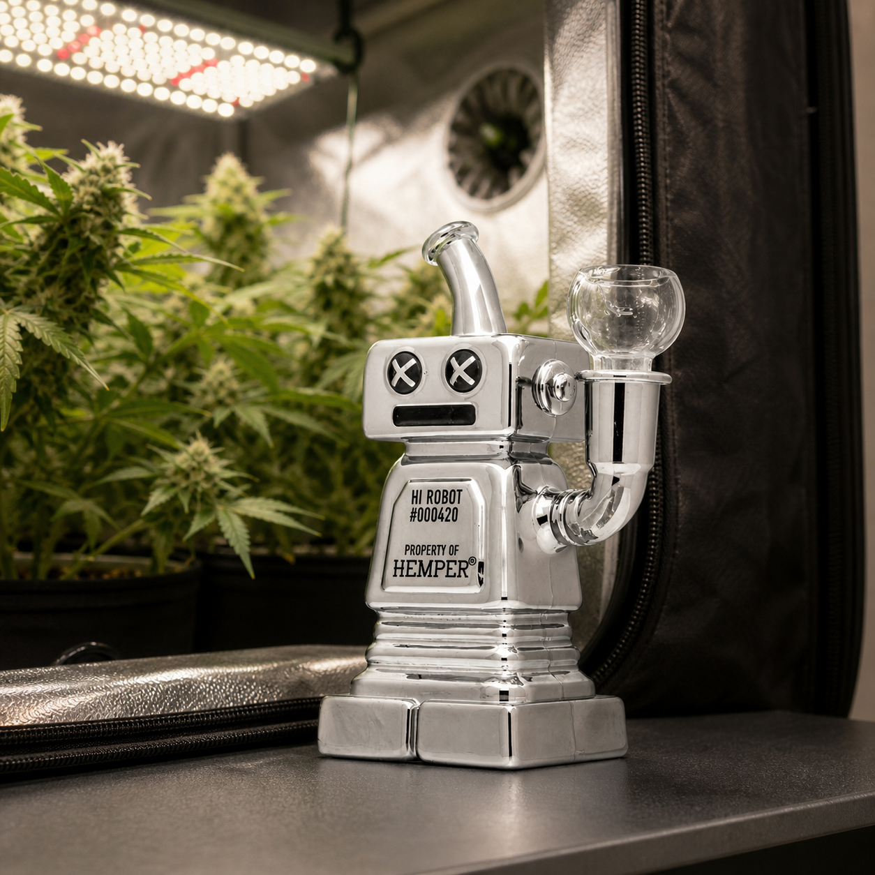 Bong Hemper Hi-Robot plateado con forma de robot, ojos en cruz y cazoleta lateral, junto a un armario con plantas y luz LED.