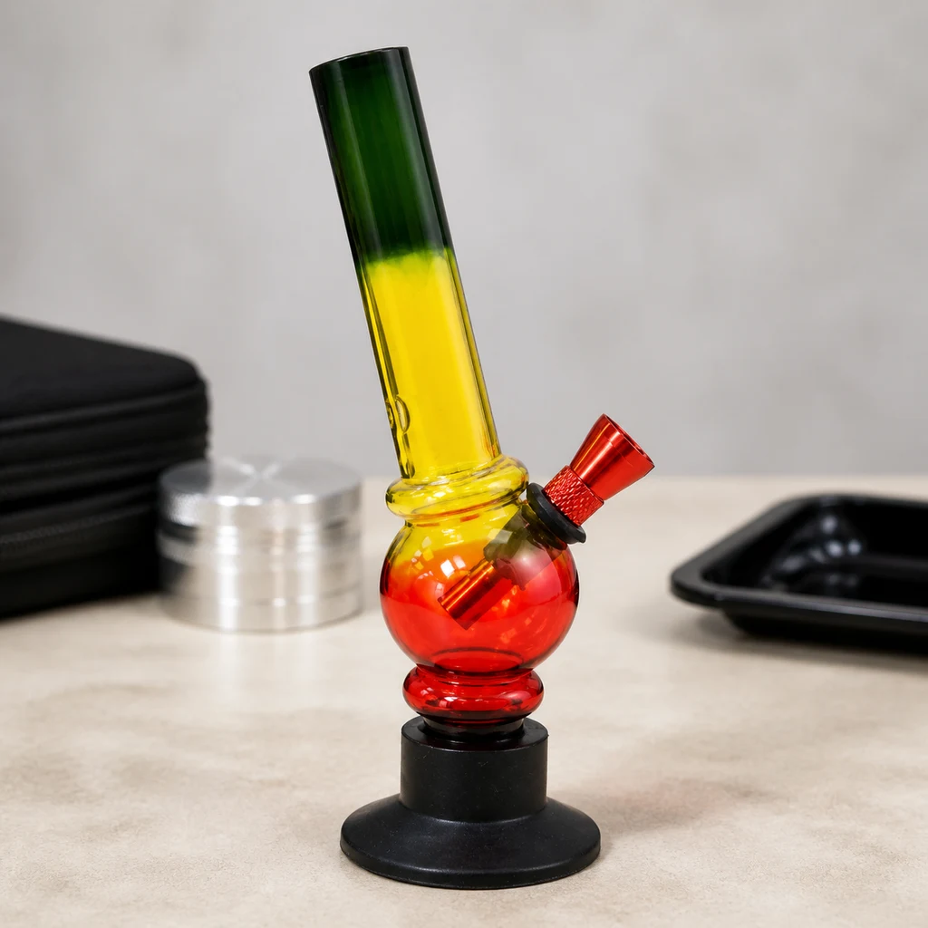 Mini bong Rasta Bubble con tubo verde y amarillo, cámara roja, cazoleta lateral y base negra sobre una mesa con accesorios.