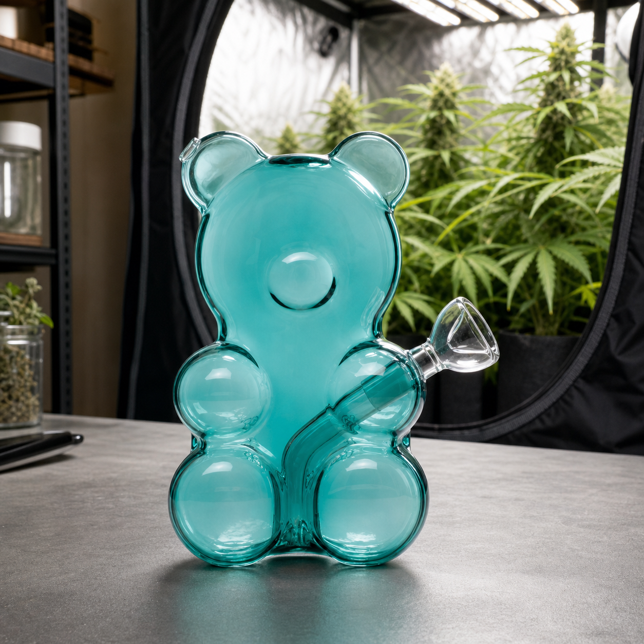 Bong Gummy Bear turquesa translúcido con forma de osito y cazoleta lateral, sobre una mesa frente a un armario con plantas.