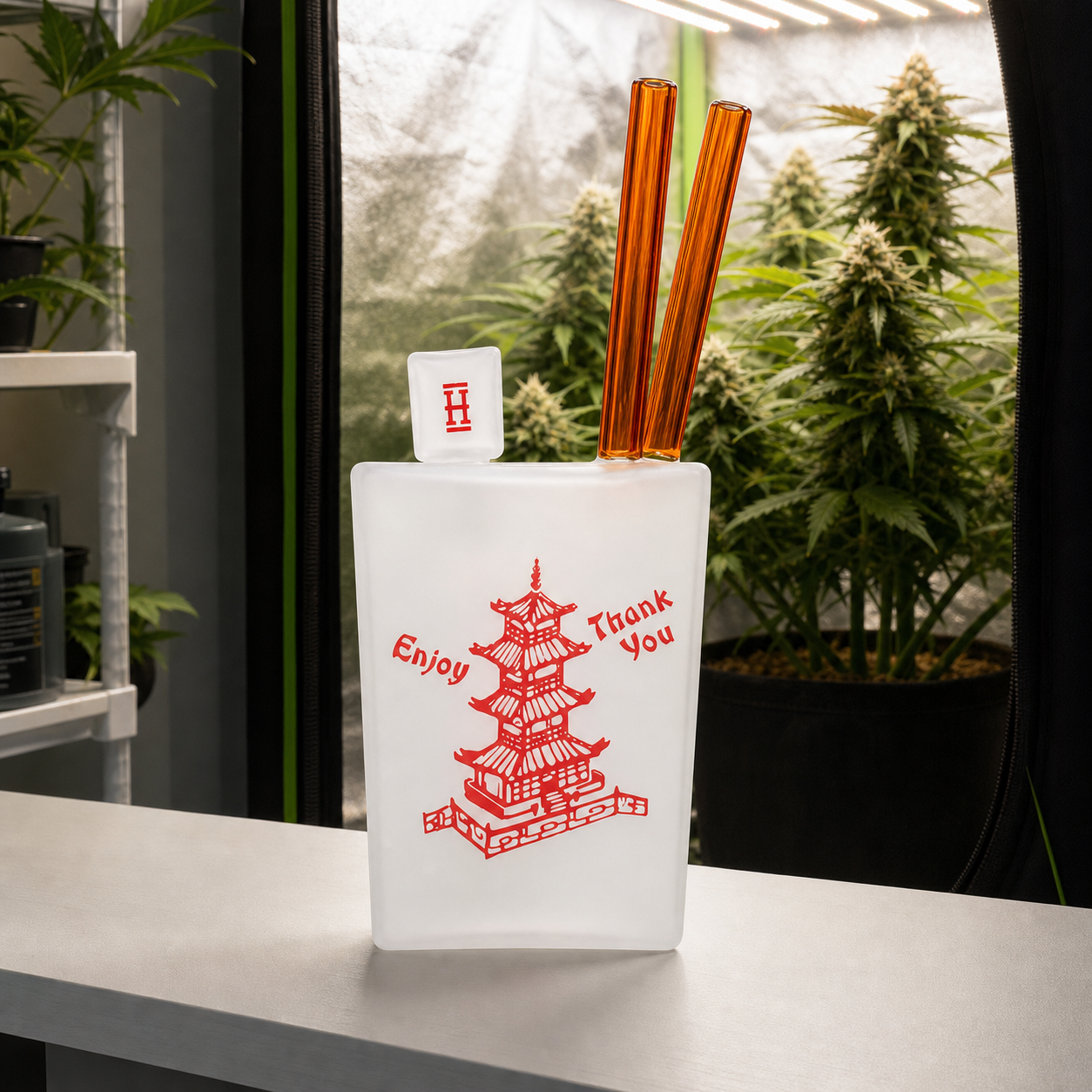 Bong Chinese Takeout XL de Hemper, con cuerpo blanco, pagoda roja y tubos ámbar, sobre una mesa frente a plantas de cannabis.