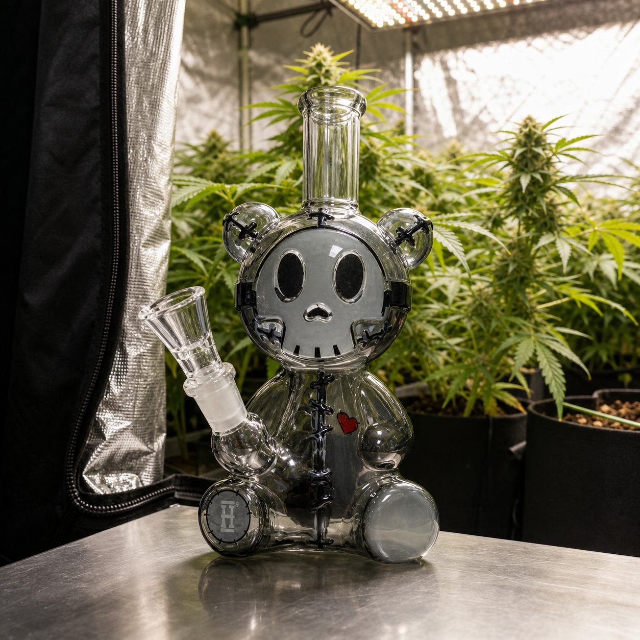 Bong transparente con forma de oso, rostro de calavera y pequeño corazón rojo, sobre una mesa ante plantas en un armario de cultivo.