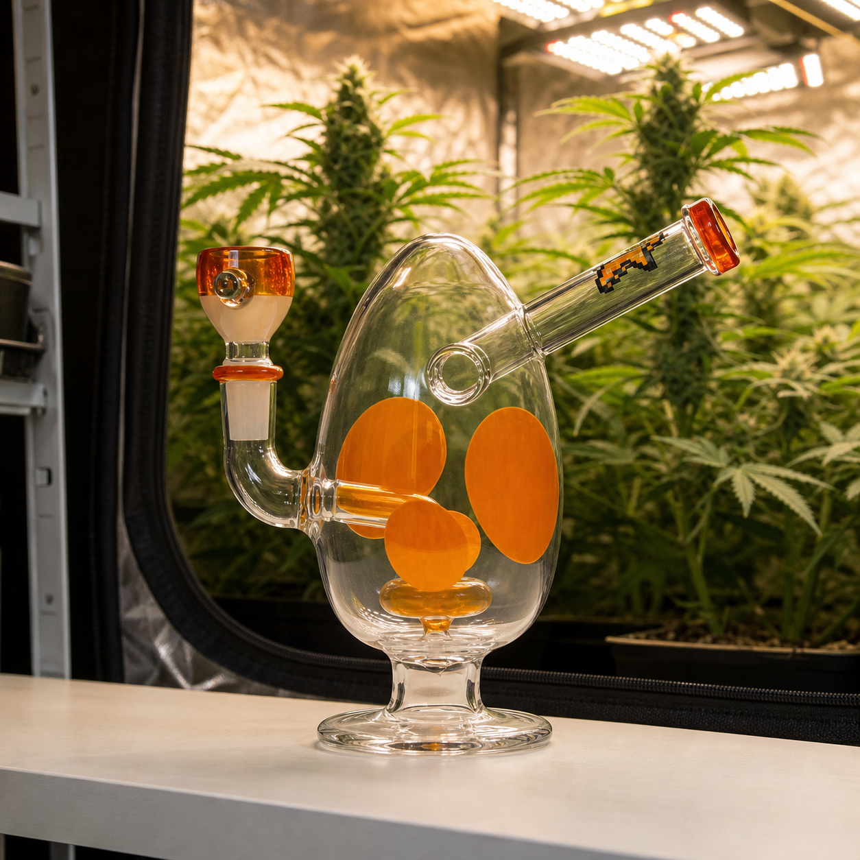 Bong Hemper transparente con cuerpo ovalado y manchas naranjas, cazoleta lateral y boquilla inclinada, ante plantas de cannabis.