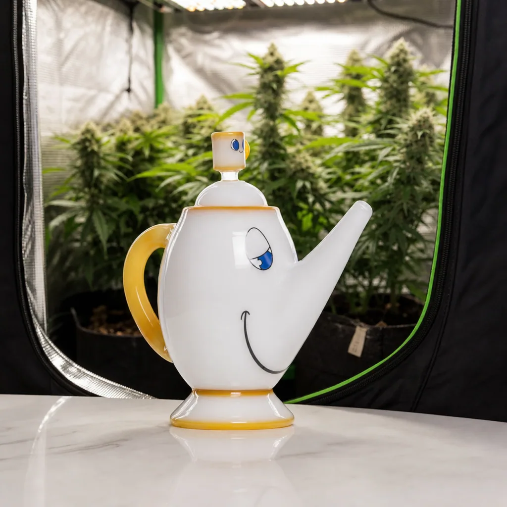 Bong Hemper Tea Pot XL con forma de tetera blanca, rostro sonriente y asa amarilla, delante de un armario con plantas.
