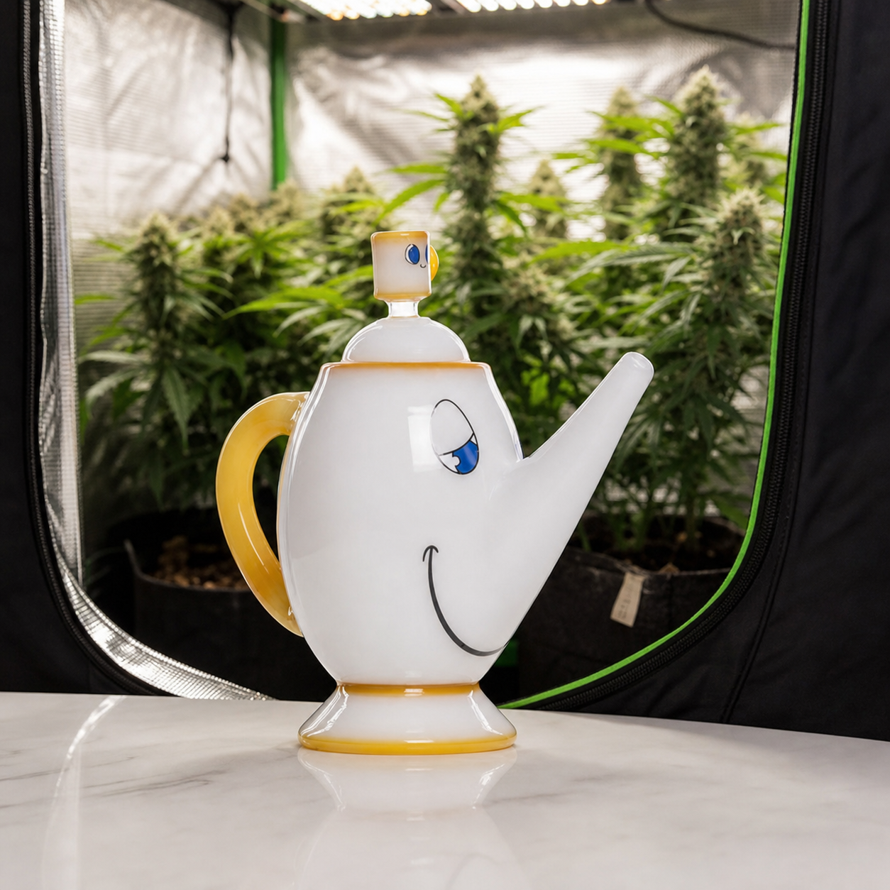 Bong Hemper Tea Pot XL con forma de tetera blanca, rostro sonriente y asa amarilla, delante de un armario con plantas.
