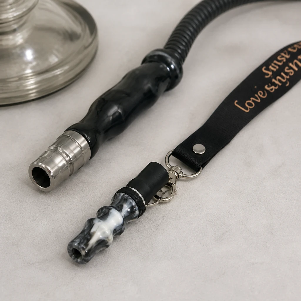 Boquilla para cachimba con acabado veteado y cordón negro con mosquetón, junto al extremo de una manguera sobre una superficie clara. Boquilla para cachimba con acabado veteado y cordón negro con mosquetón, junto al extremo de una manguera sobre una superficie clara.