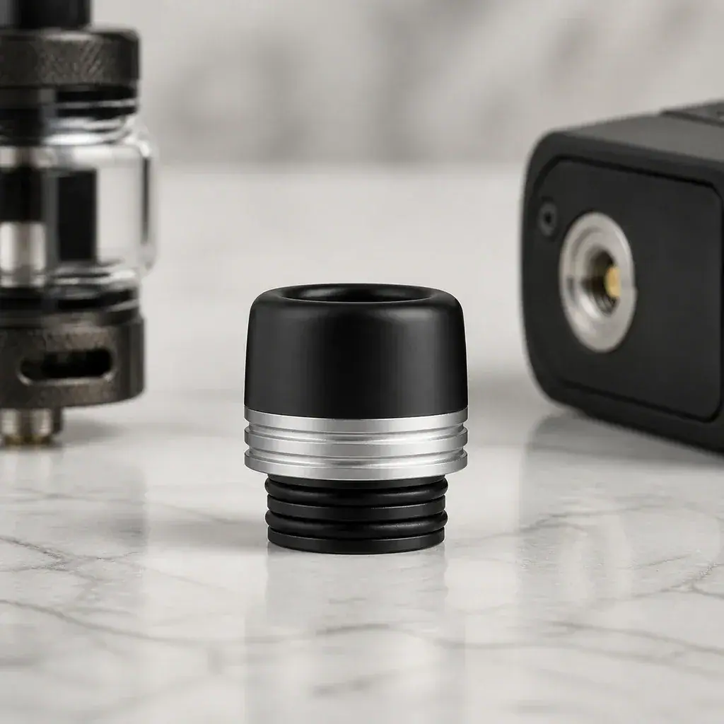 Boquilla Voopoo Drip Tip 810 negra con aro plateado y juntas negras, sobre una mesa con un depósito y un dispositivo desenfocados detrás.
