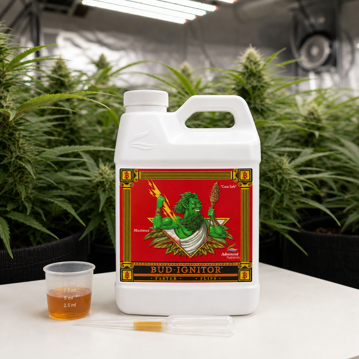 Garrafa blanca de Bud Ignitor de Advanced Nutrients con etiqueta roja, vaso dosificador y pipetas ante plantas en floración.