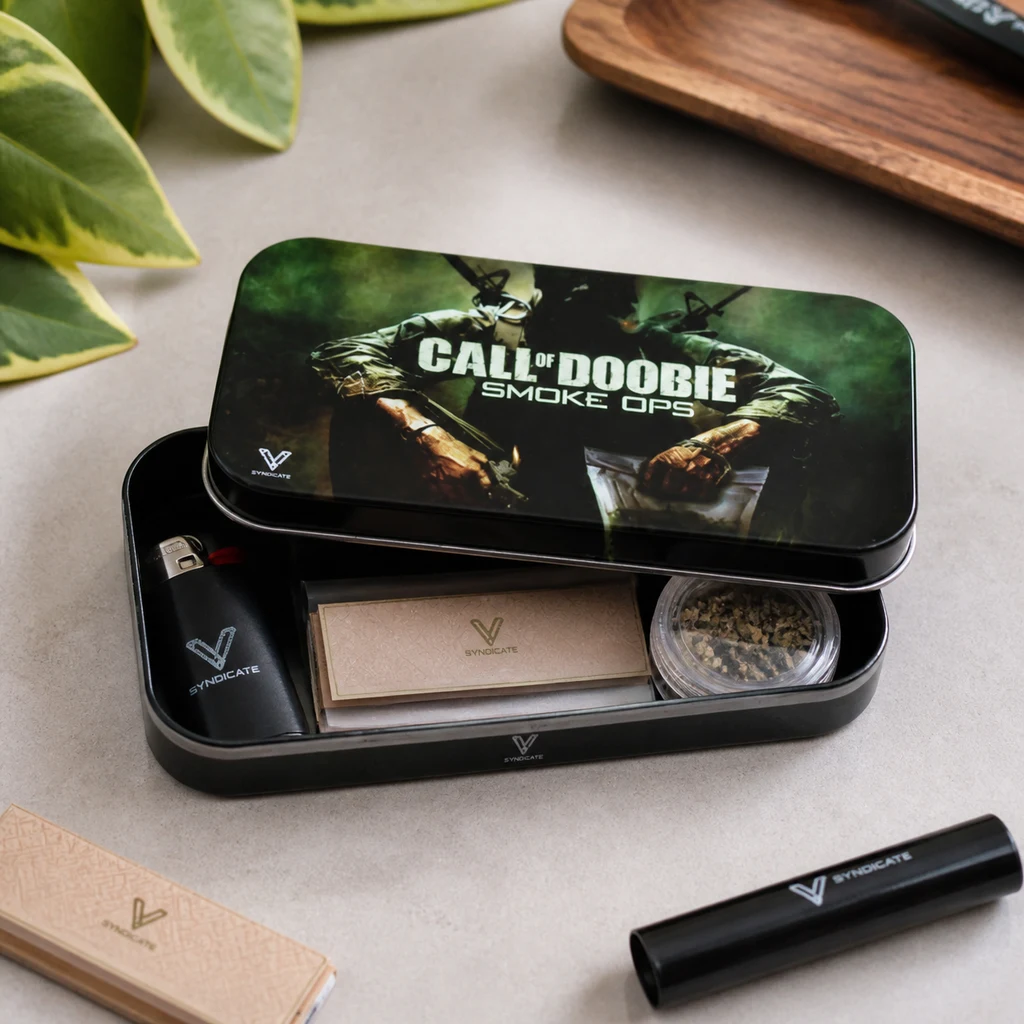 Caja metálica Call of Doobie abierta, con soldados en la tapa y accesorios para liar dentro; un tubo negro descansa al lado. Caja metálica Call of Doobie abierta, con soldados en la tapa y accesorios para liar dentro; un tubo negro descansa al lado.