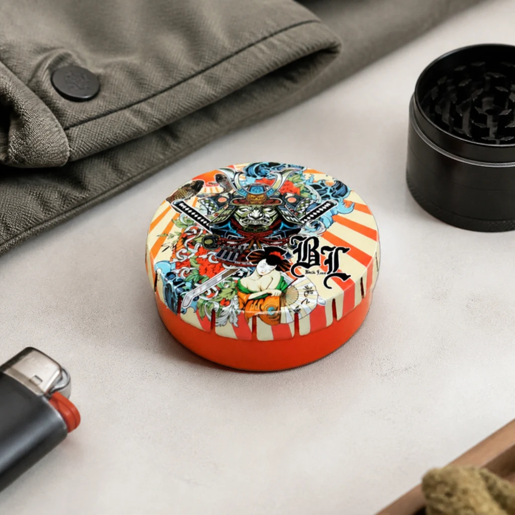 Caja redonda Ronin & Geisha con base naranja y tapa ilustrada con figuras japonesas, sobre una mesa junto a un grinder negro.