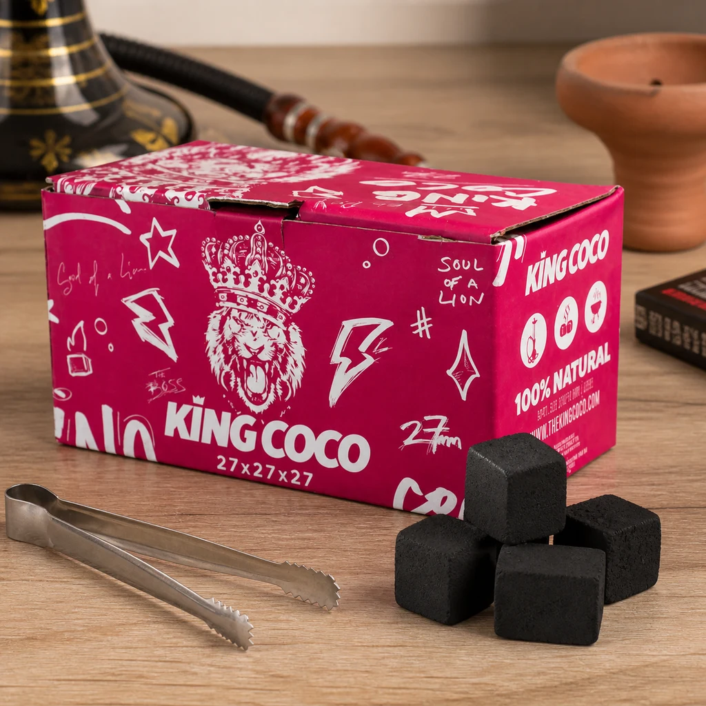 Caja rosa de carbón King Coco junto a varios cubos negros y unas pinzas metálicas sobre una mesa, con una cachimba al fondo.