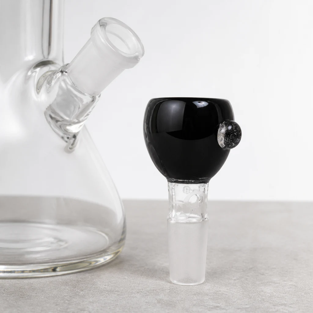 Cazoleta negra de cristal con saliente lateral y conexión transparente esmerilada, colocada junto a un bong de cristal transparente.