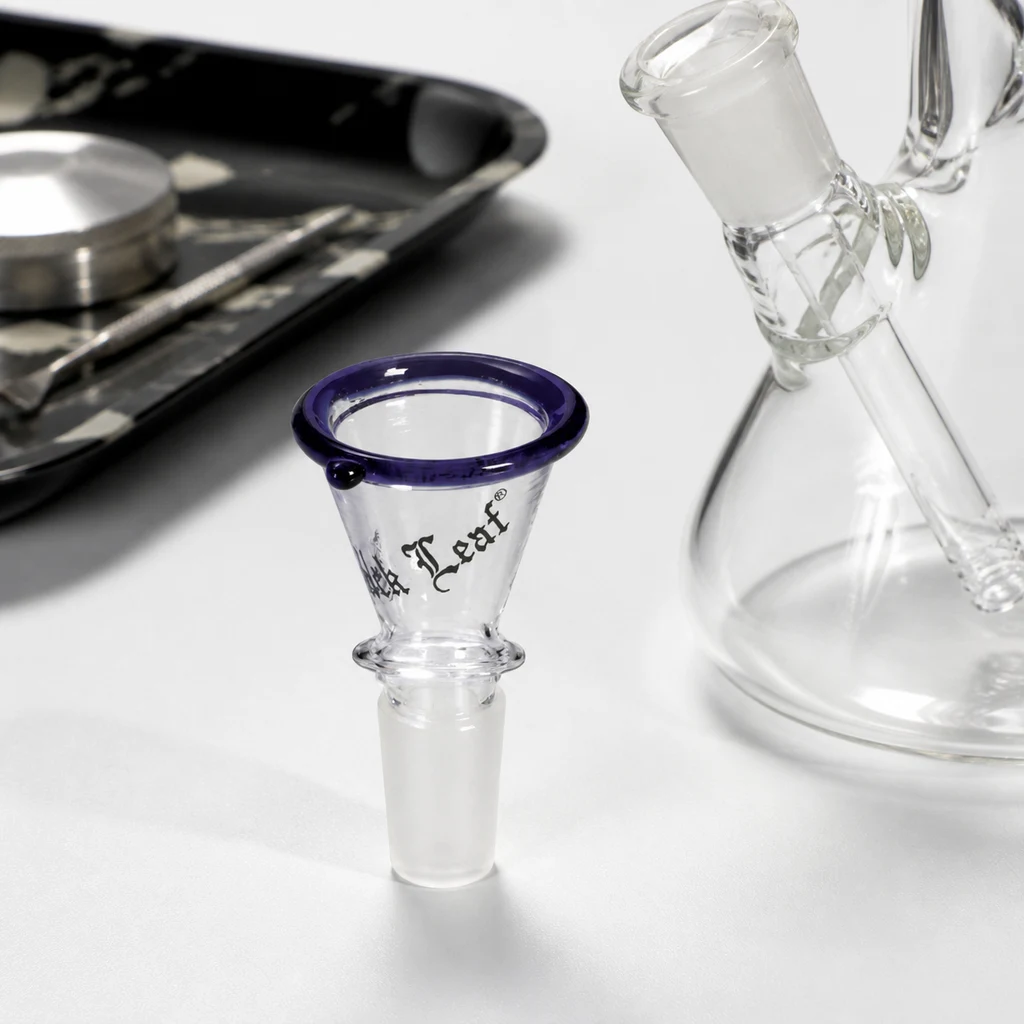 Boquilla cónica de cristal Black Leaf con borde violeta y conexión esmerilada, junto a un bong transparente y una bandeja desenfocada.
