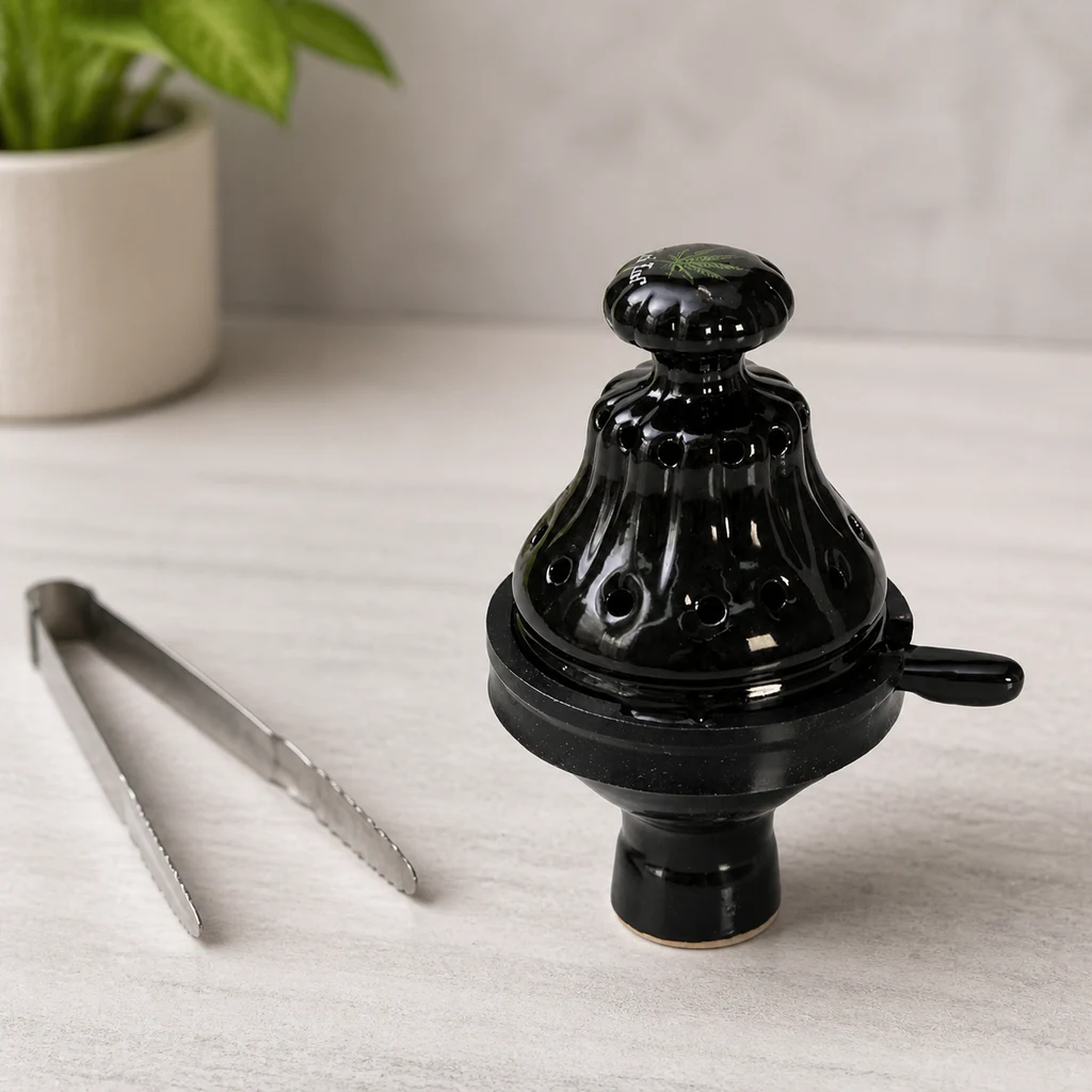 Cazoleta de cachimba negra con tapa perforada, pomo y asa lateral sobre una mesa clara, junto a unas pinzas metálicas.