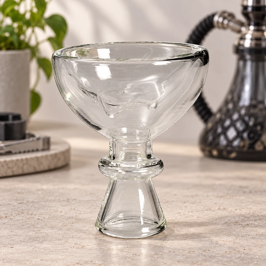 Cuenco de cachimba de vidrio transparente, con copa ancha y base cónica, sobre una mesa con una cachimba desenfocada al fondo.