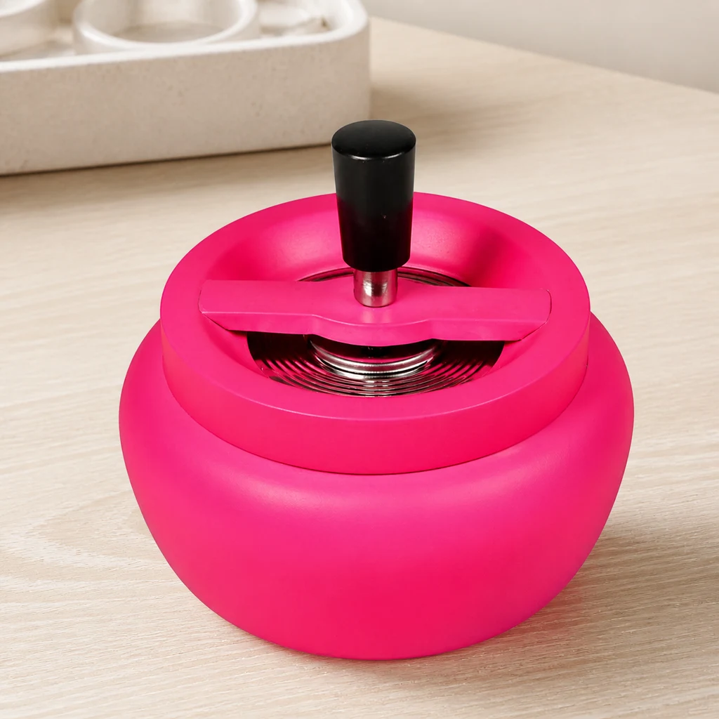 Cenicero rosa de cuerpo redondeado, con pulsador negro central y mecanismo plateado visible, sobre una mesa clara en una escena generada.