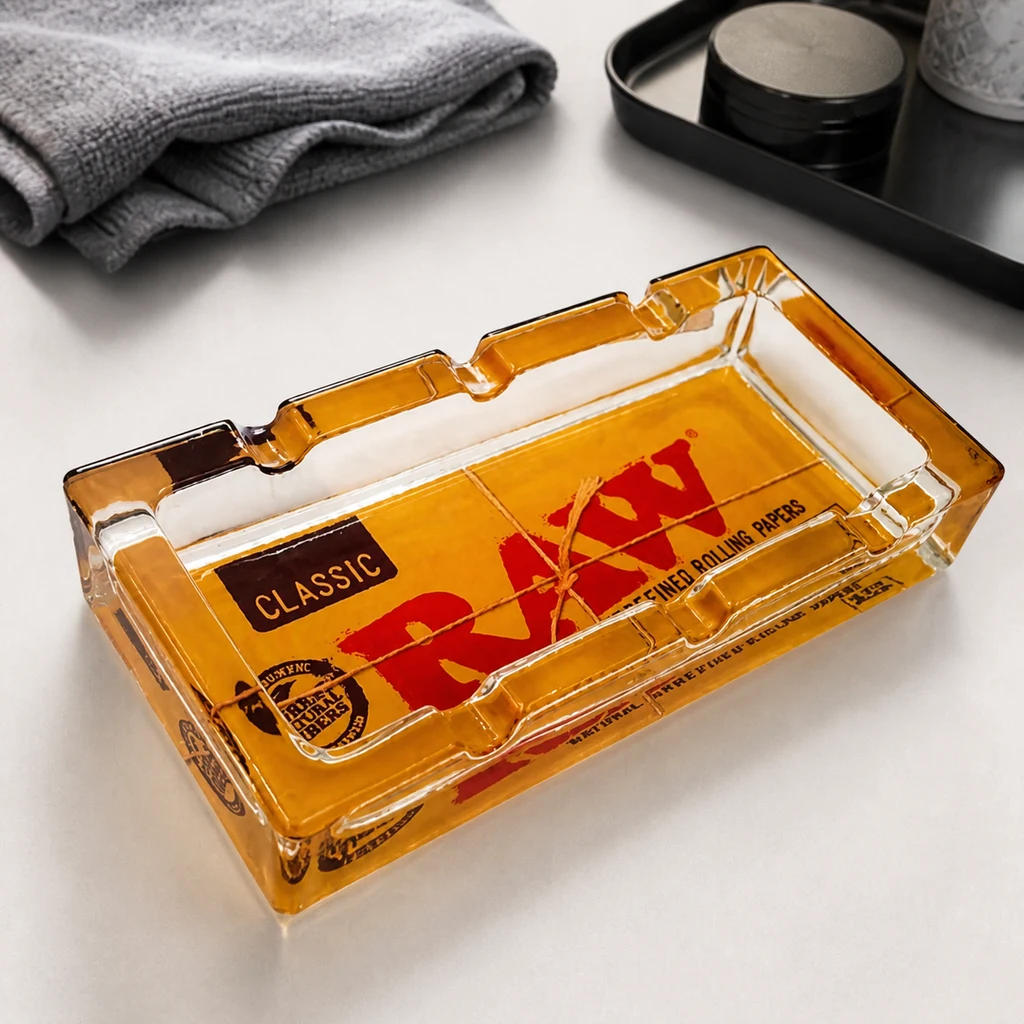 Cenicero rectangular RAW de cristal ámbar, con muescas en los bordes y logotipo rojo, sobre una mesa junto a una toalla gris.