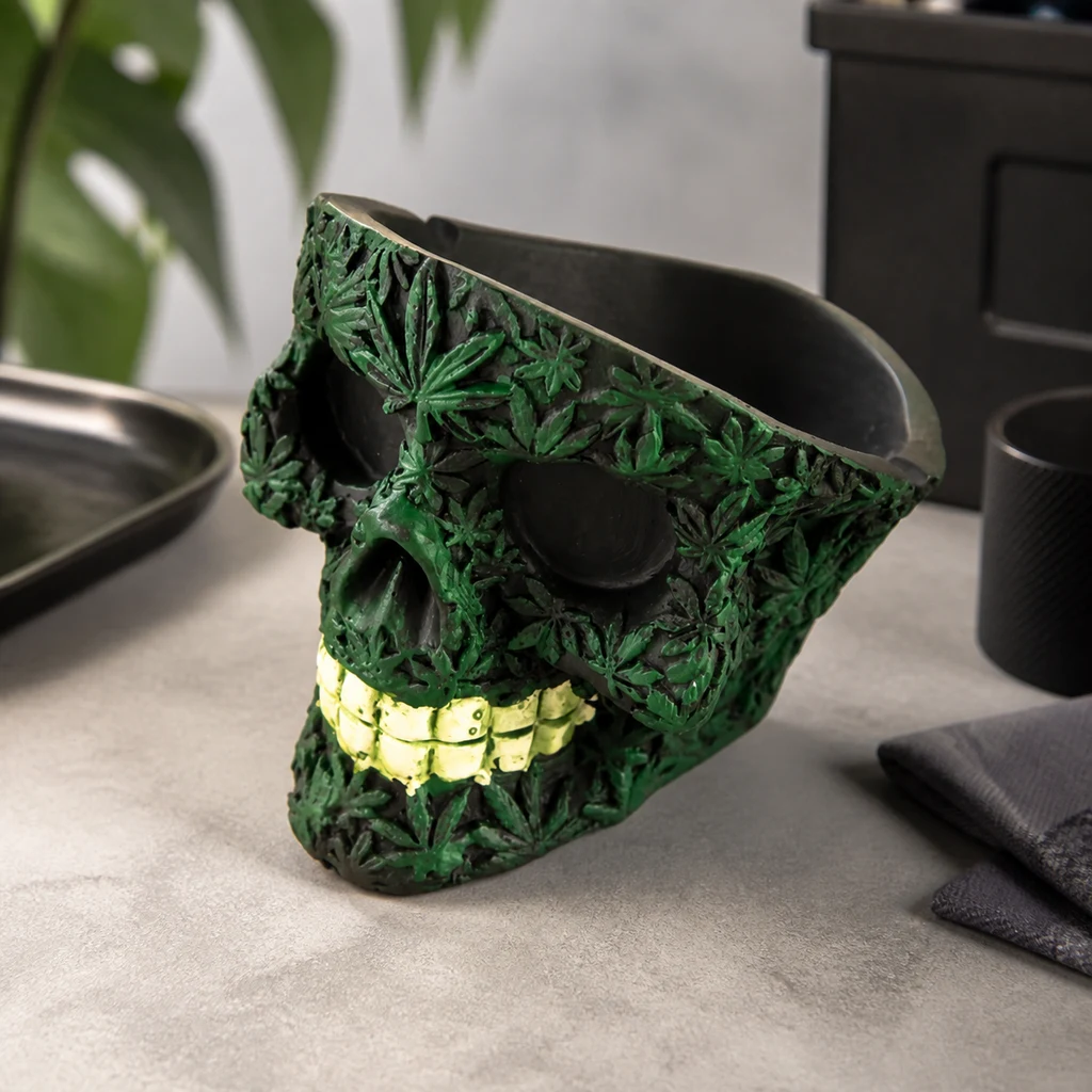 Cenicero con forma de calavera, hojas verdes en relieve y dientes dorados, con recipiente abierto en la parte superior, sobre una mesa.