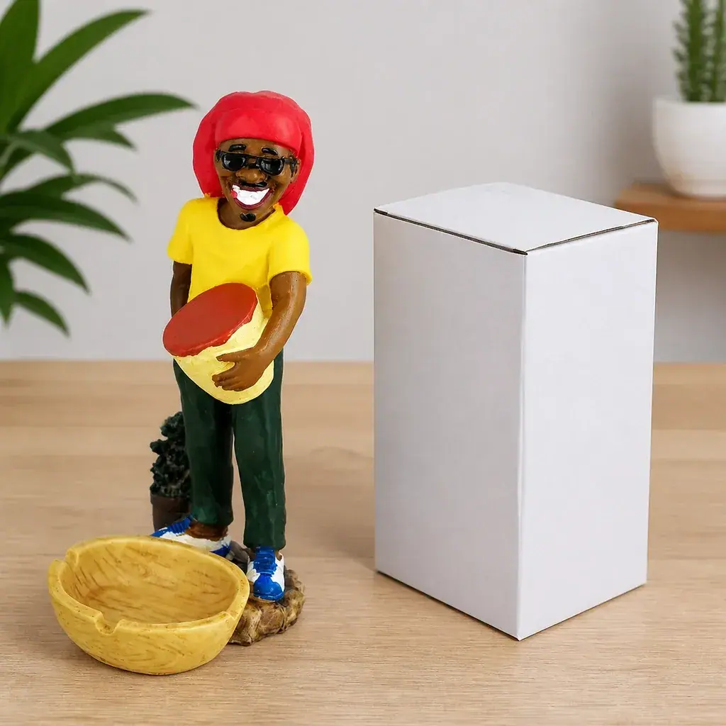 Cenicero Smiling Rasta con figura sonriente, gafas oscuras, gorro rojo y camiseta amarilla, junto a una caja blanca sobre una mesa.