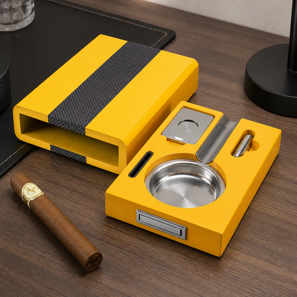 Cenicero amarillo para puros con cuenco plateado, cortador y punzón, junto a una cubierta con franja oscura y un puro sobre la mesa.