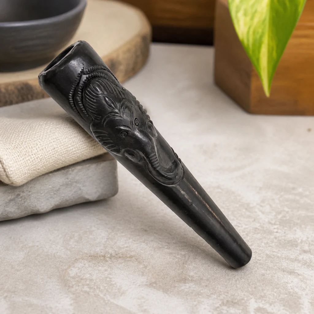 Chillum negro de forma cónica con una cabeza de elefante en relieve, apoyado en diagonal sobre una tela y una base gris. Chillum negro de forma cónica con una cabeza de elefante en relieve, apoyado en diagonal sobre una tela y una base gris.