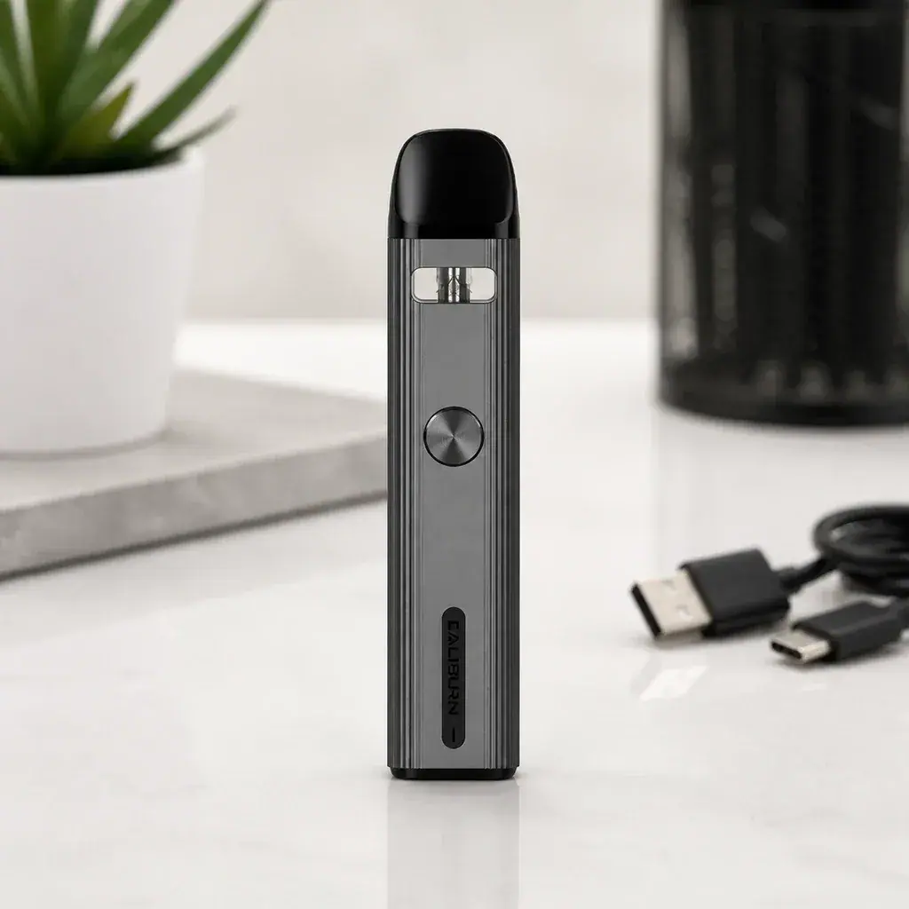 Uwell Caliburn G2 gris oscuro, de pie sobre una mesa, con botón circular, boquilla negra y cable USB al lado.