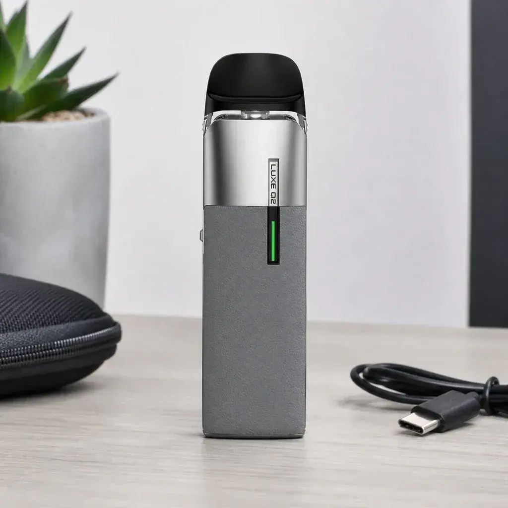 Escena generada del Vaporesso Luxe Q2 gris con boquilla negra e indicador verde sobre una mesa, junto a un cable USB y un estuche.
