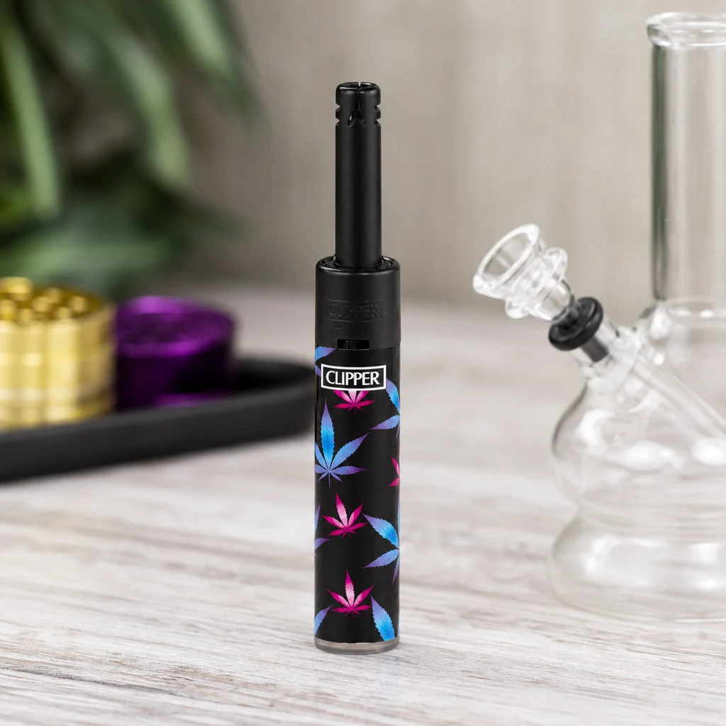 Mechero Clipper Minitube negro con tubo alargado y hojas azules y rosas, de pie junto a un bong de cristal y grinders desenfocados.