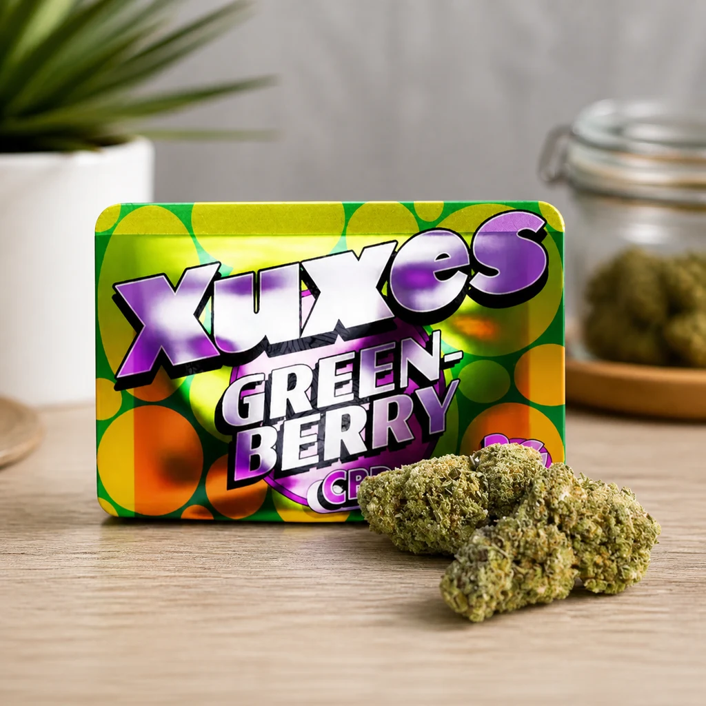 Envase verde y amarillo Xuxes Green Berry con letras moradas, junto a varios cogollos sobre una mesa de madera.
