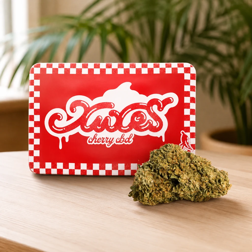 Cogollo junto a una caja roja de Xuxes Cherry CBD con bordes de cuadros blancos, sobre una mesa y con plantas desenfocadas al fondo.