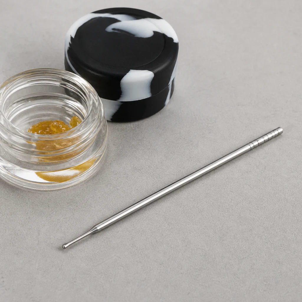 Dabber BHO plateado en diagonal junto a un tarro abierto con sustancia ámbar y un recipiente negro y blanco sobre una superficie gris.