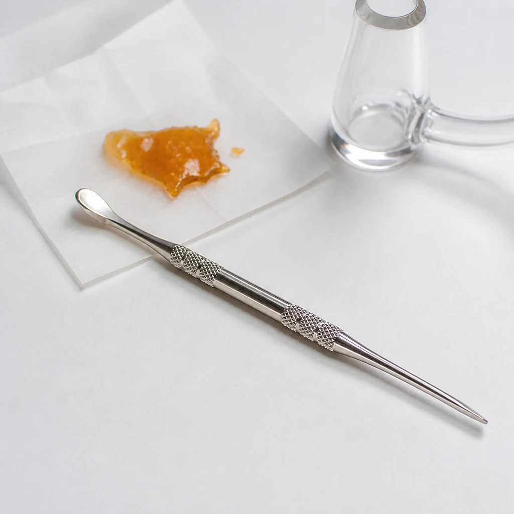 Dabber plateado en diagonal, con zonas de agarre texturizadas, punta fina y extremo de cuchara, junto a un concentrado ámbar y vidrio.