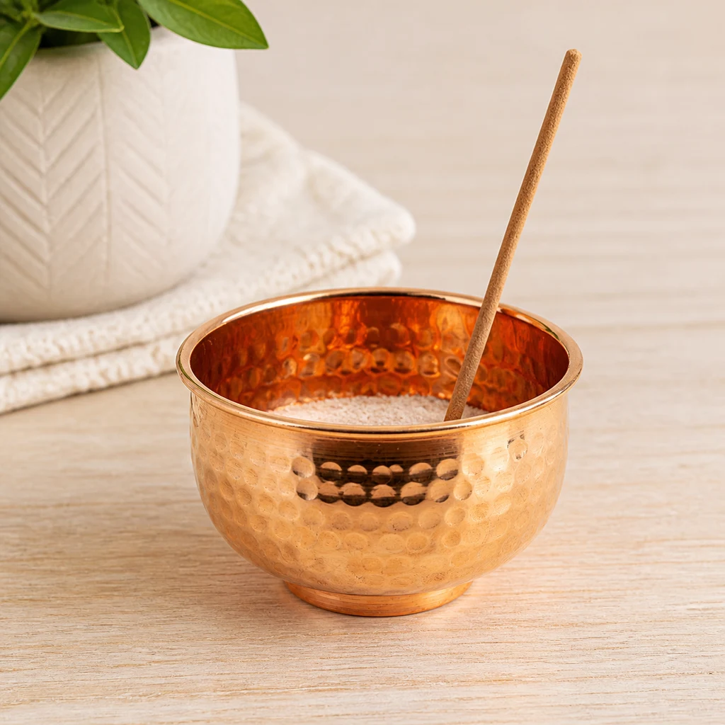 Cenicero de incienso Dhupa en forma de cuenco cobrizo martillado, con relleno claro y una varilla inclinada sobre una mesa de madera. Cenicero de incienso Dhupa en forma de cuenco cobrizo martillado, con relleno claro y una varilla inclinada sobre una mesa de madera.