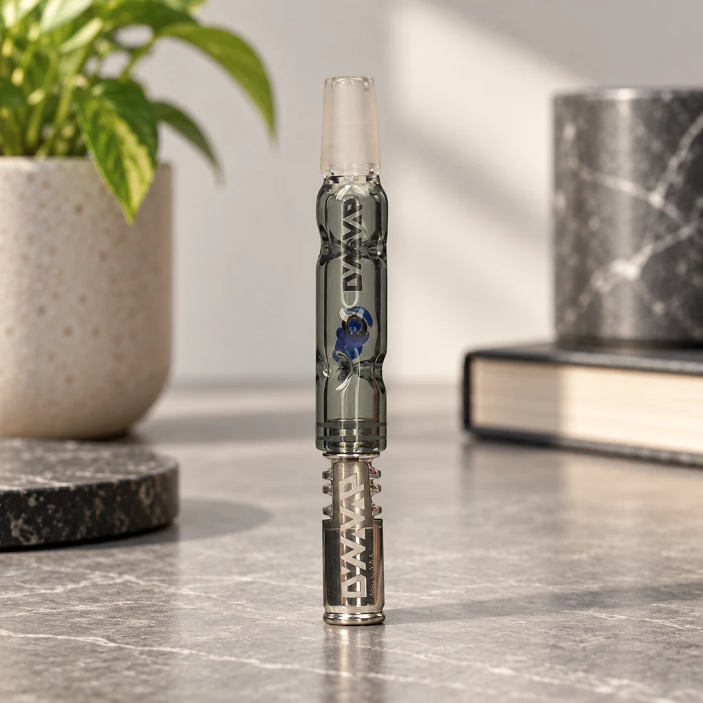 DynaVap BB3 Gris en posición vertical, con cuerpo translúcido, detalles azules y extremo metálico, sobre una mesa junto a una planta y libros.