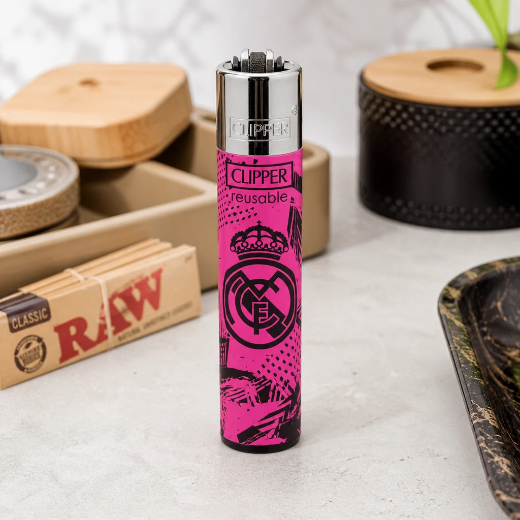 Escena generada con mechero Clipper rosa y negro con escudo del Real Madrid, de pie junto a papel RAW y accesorios sobre una mesa.