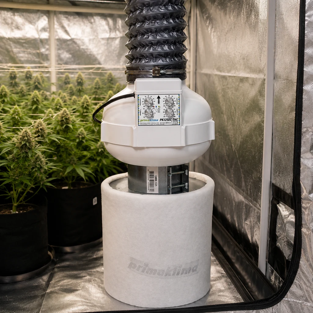 Extractor Prima Klima con controles frontales, conectado a un conducto negro y un filtro cilíndrico blanco en un armario con plantas.