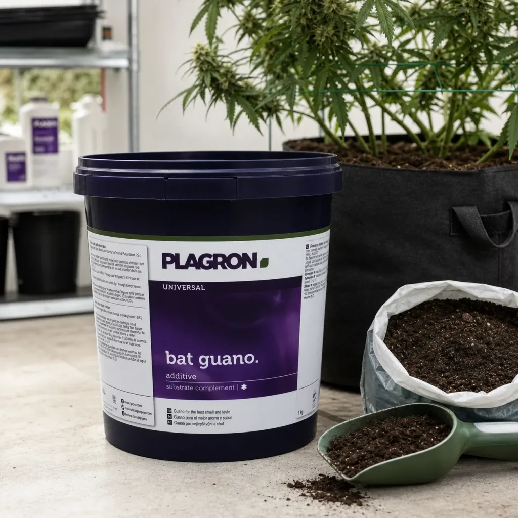 Cubo de guano de murciélago Plagron junto a una bolsa abierta y una pala con sustrato, delante de una planta en maceta textil.