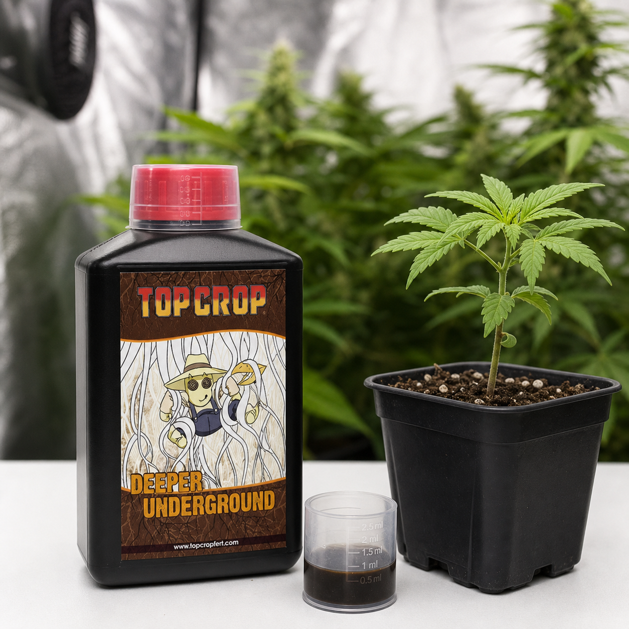 Botella de Deeper Underground Top Crop junto a un vaso dosificador con líquido oscuro y una planta en maceta, en un cultivo interior.