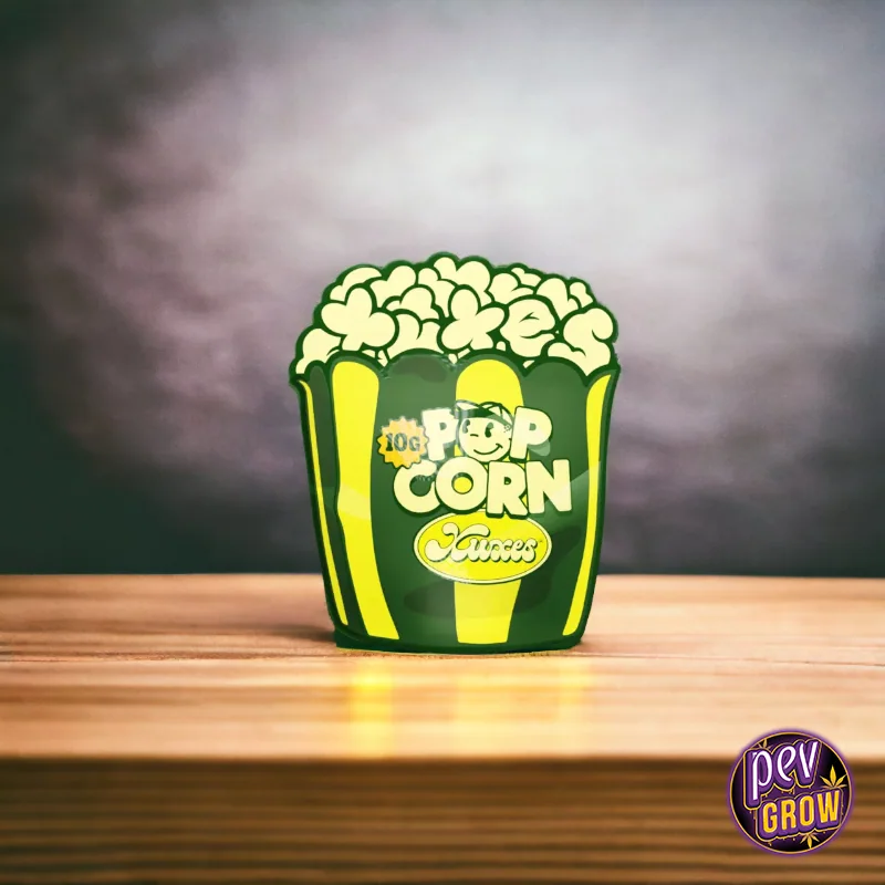 Envase de Pop Corn Xuxes con diseño verde y amarillo en forma de caja de palomitas, marcado 10G, sobre una mesa de madera.