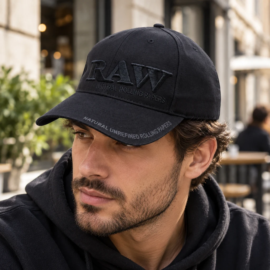 Hombre con gorra negra RAW de visera curva y logotipo bordado en negro, en primer plano con una terraza desenfocada al fondo.