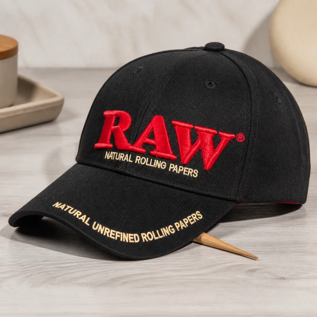Gorra negra con logotipo RAW bordado en rojo y letras beige en el frontal y la visera, apoyada sobre una superficie clara.