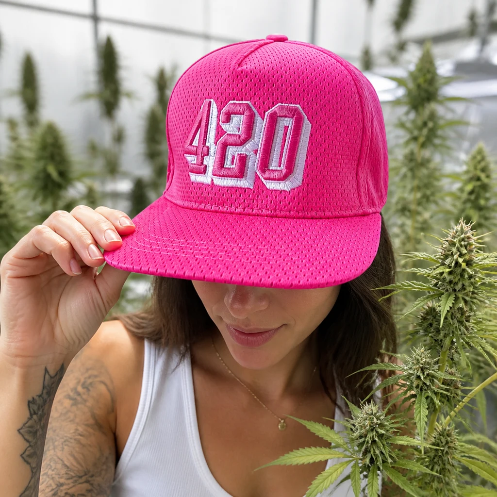 Mujer sujetando la visera de una gorra rosa con «420» bordado en relieve, entre plantas de cannabis y con los ojos ocultos por la visera.