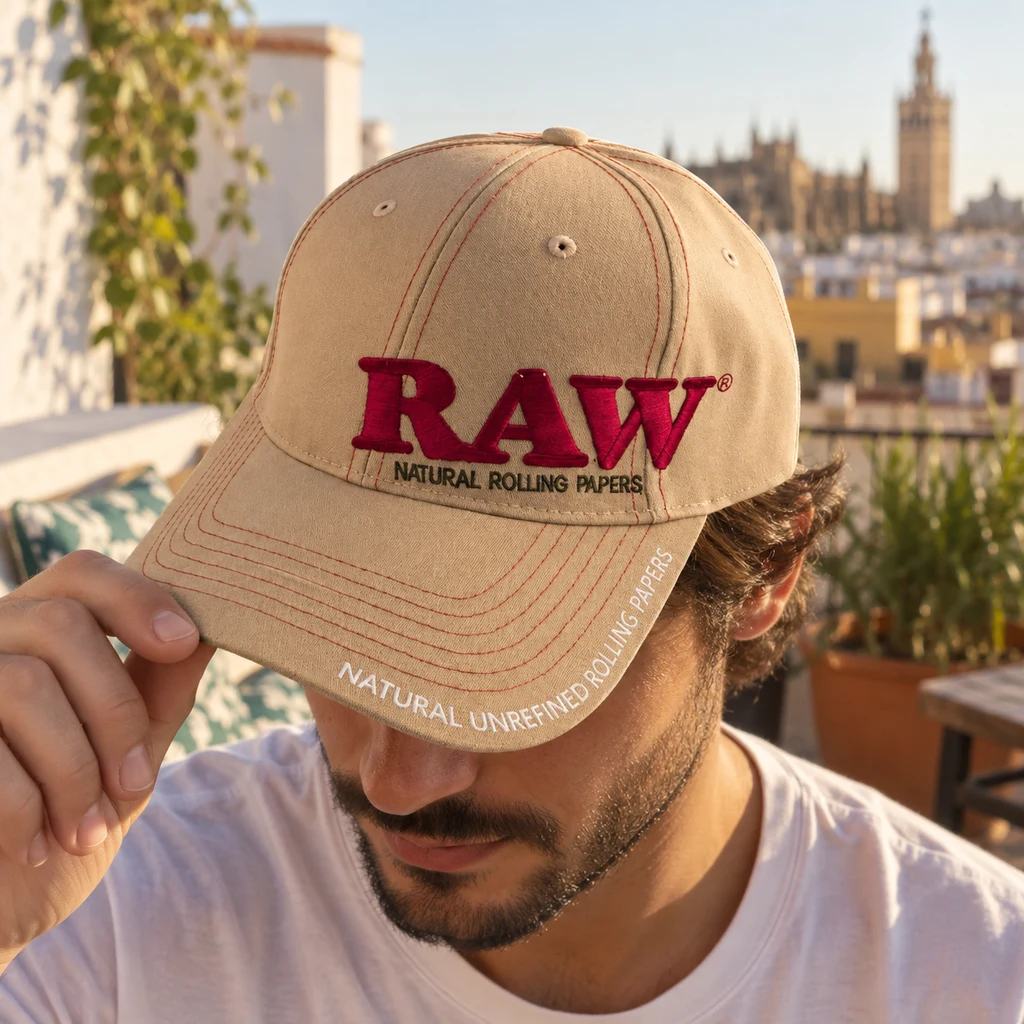Un hombre sujeta la visera de una gorra beige con RAW bordado en rojo y costuras rojas, en una terraza con edificios al fondo.