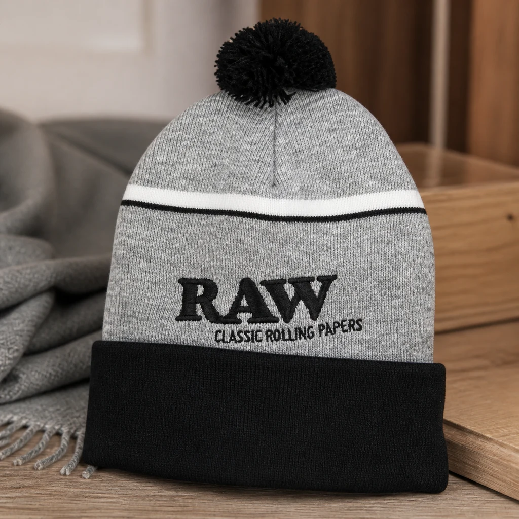 Gorro RAW gris con pompón negro, vuelta negra y franjas blanca y negra, colocado sobre una superficie de madera. Gorro RAW gris con pompón negro, vuelta negra y franjas blanca y negra, colocado sobre una superficie de madera.