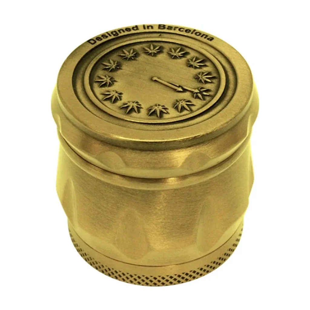 Grinder dorado con relieve de reloj y hojas de cannabis en la tapa, inscripción «Designed in Barcelona» y base con textura romboidal.