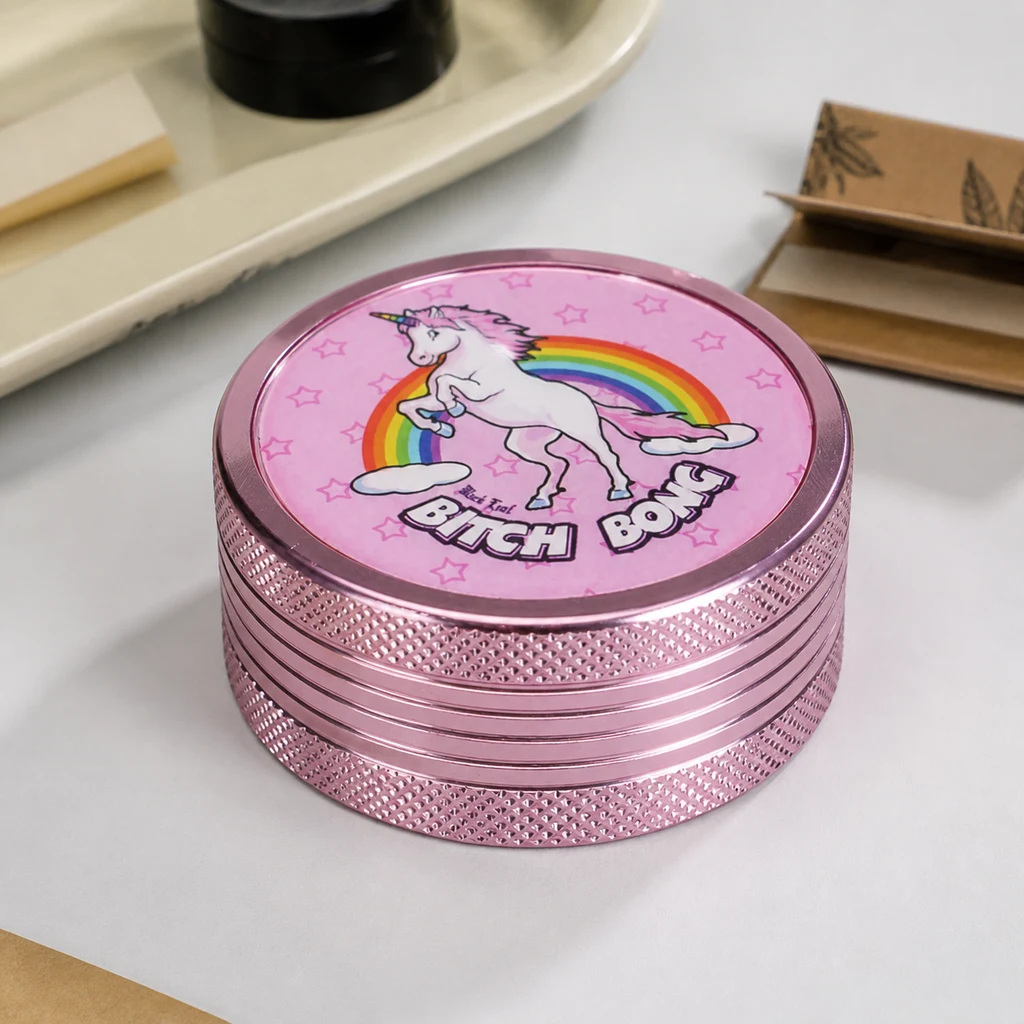 Grinder rosa Bitch Bong cerrado, con bordes texturizados y un unicornio ante un arcoíris en la tapa, sobre una mesa clara.