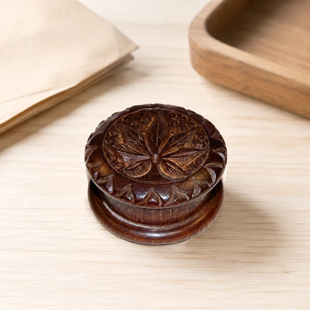 Molinillo de madera Palosanto cerrado, de tono marrón oscuro, con una hoja de cannabis tallada en la tapa sobre una mesa clara.