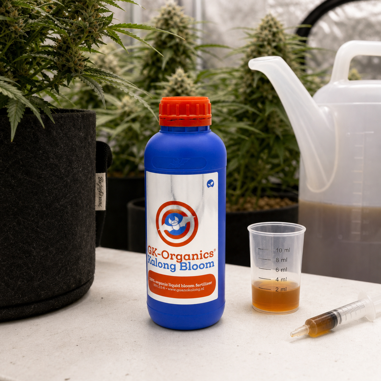 Botella azul de GK-Organics Kalong Bloom junto a un vaso graduado con líquido ámbar, una jeringa y una regadera, con plantas al fondo.