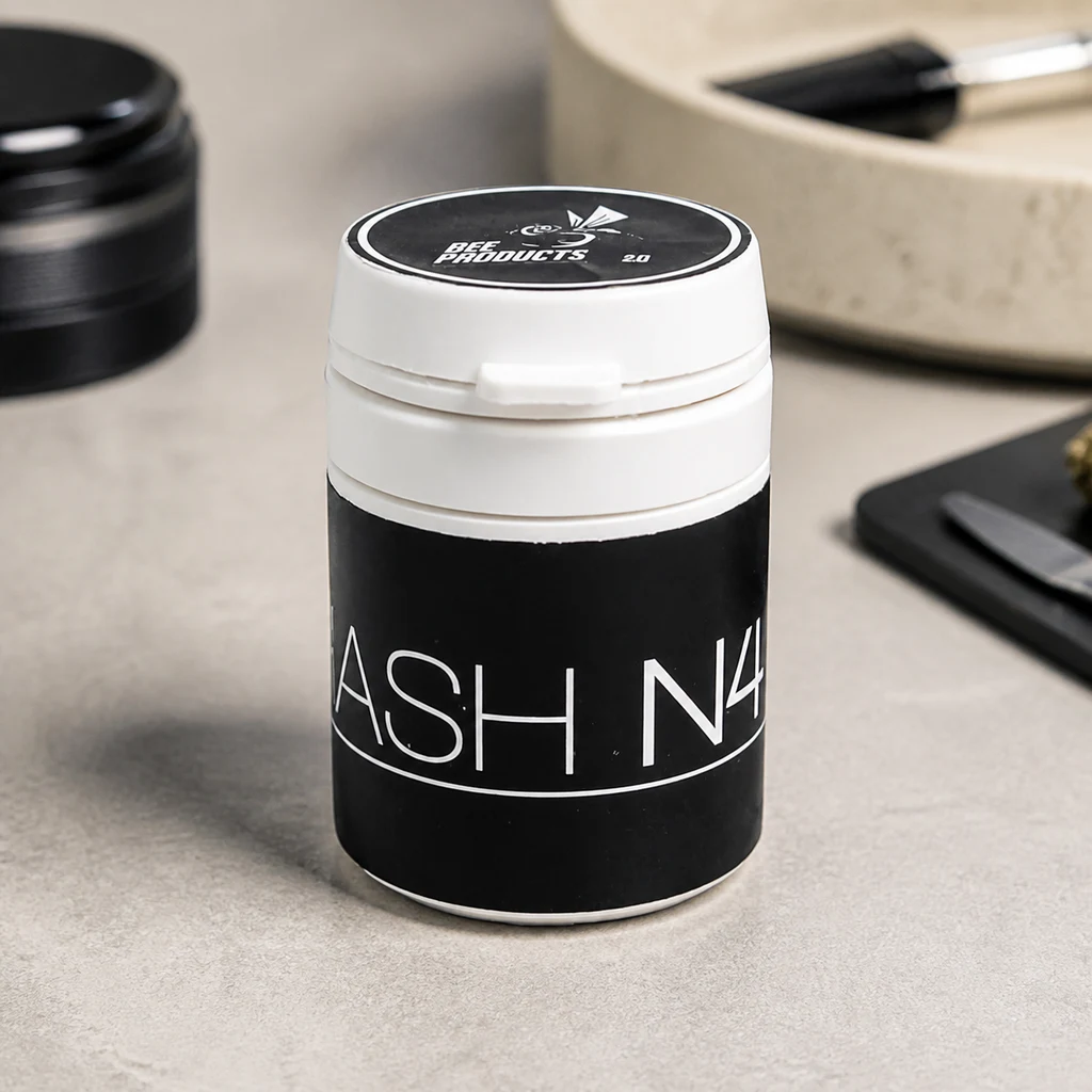 Bote blanco de Hash Dry N4 CBD Bee con etiqueta negra sobre una mesa, junto a un grinder y una bandeja desenfocados.